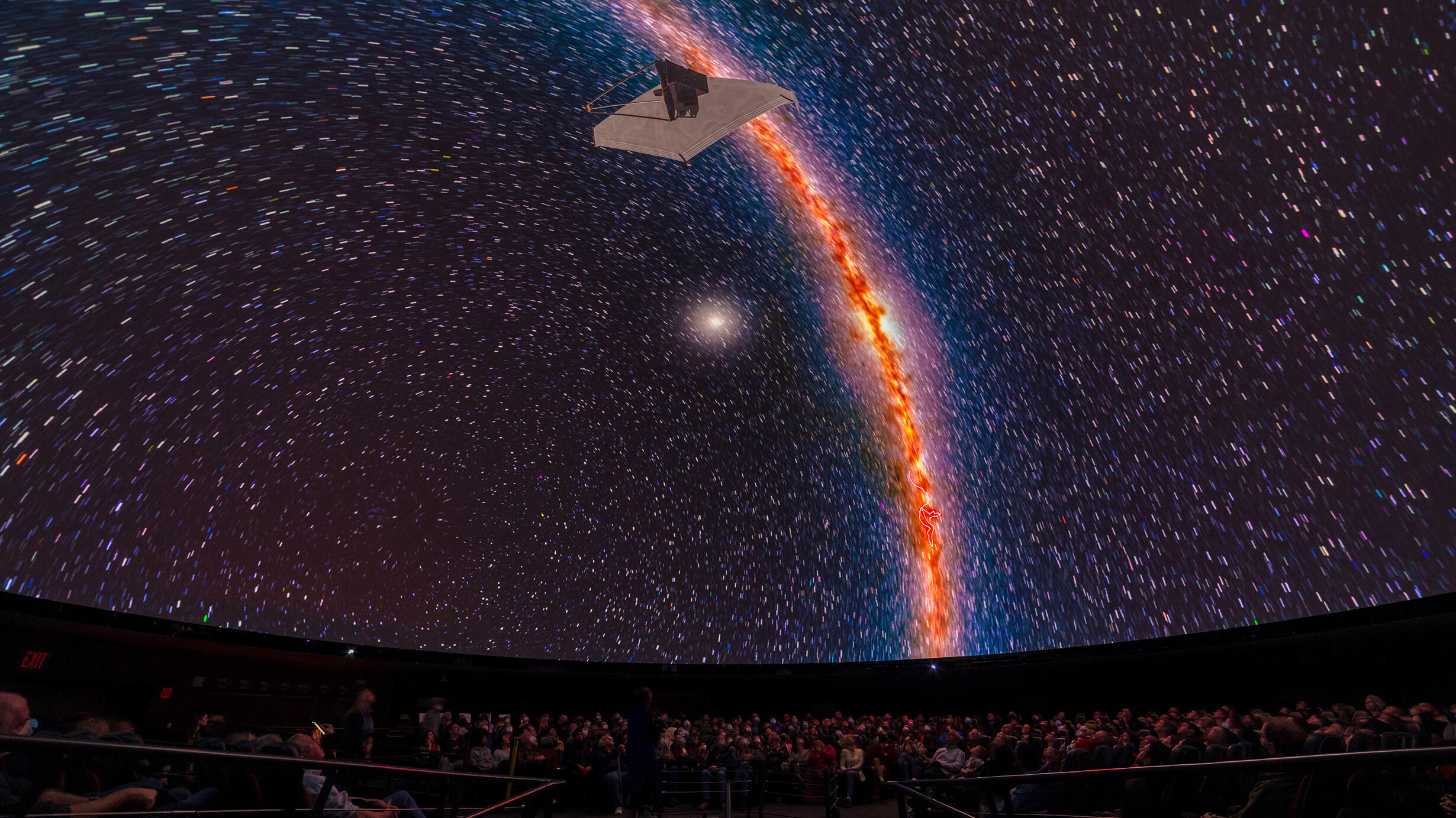 A wide-angle photograph of an audience inside the Hayden Planetarium, looking up at a depiction of a spacecraft against a backdrop of white stars.