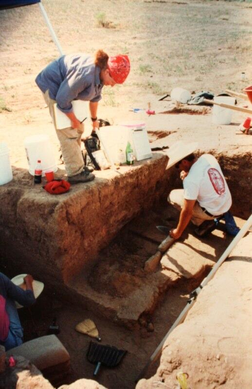 Two people excavating what looks like a low wall or building foundation in an arid dusty environment in bright sunlight.