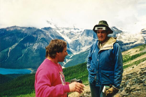 Martin Shugar (left) speaks to Des Collins (right) on a hill, with mountains visible in the background.