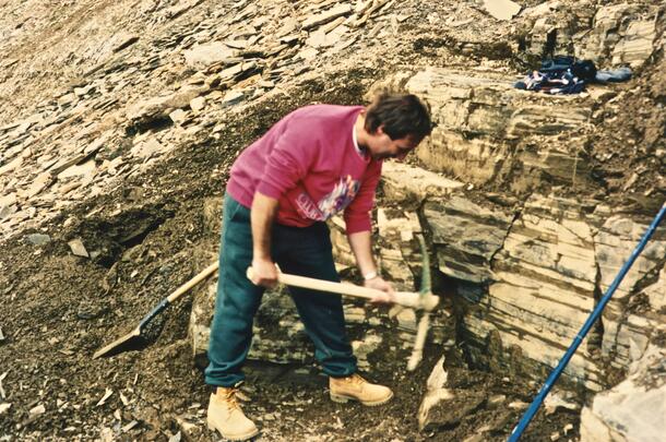Martin Shugar, holding a pick-axe, leans over to chip away at sloping rock.
