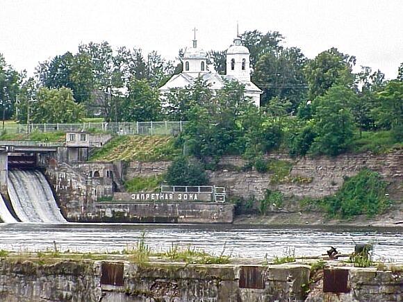 The banks of the Volkhov River with a dam visible to the left and a light-colored church above the slopes of the bank.