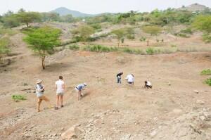 A group of six researchers outdoors in a dry, grassless setting, bending down to examine the ground.