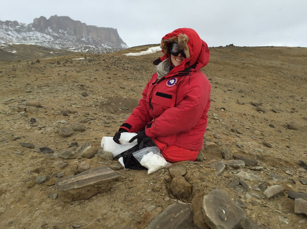 Abby West, wearing a brightly colored, hooded puffer coat and goggles, sits on dirt on a mountain.