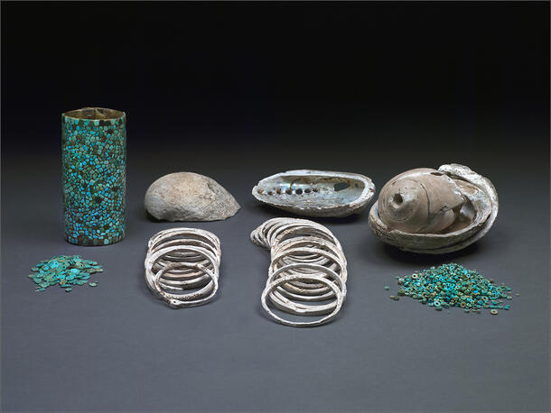 Artifacts are lined up on a studio backdrop, from left, turquoise beads, a mosaic cylinder basket, shells, shell bracelets and more beads.