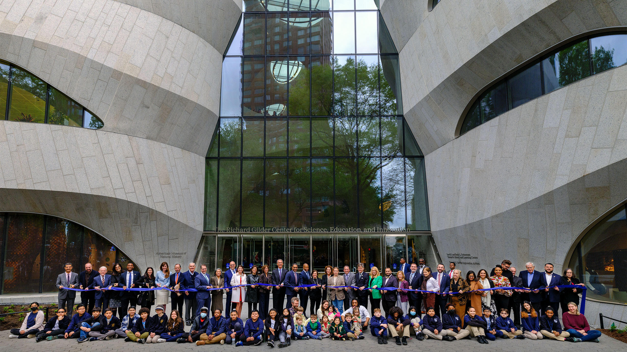 Over 70 people pose in front of the Gilder Center in two rows, with adults standing in back holding a long ribbon and children sitting in front.