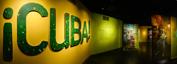 The entrance to the ¡Cuba! exhibition at the Museum, featuring a colorful sign (reading "¡Cuba!") and three life-size photos of Cuban people.
