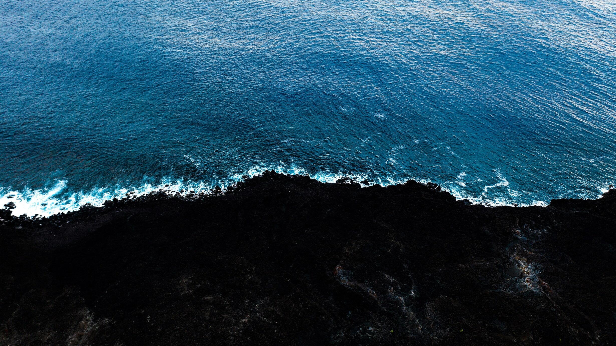 An aerial photograph of a waves on the shore: a rich blue swath of ocean meets a dark, rocky coast.
