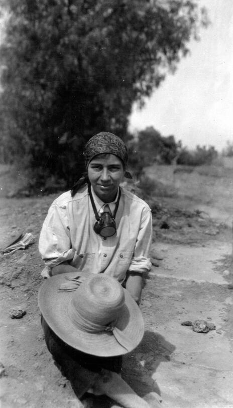 A woman, Susannah Clapp Vaillant, kneeling in the soil at an excavation site, looking at the camera, holding a wide-brimmed sun hat in her hand.