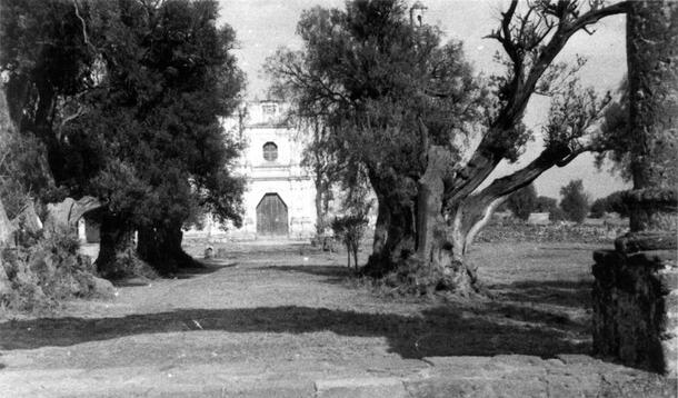 Chiconaulta Church, a light-colored building with a wide door, seen in the distance at the end of a road lined with trees.