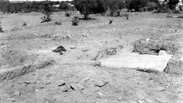 A excavation site on flat dry dusty terrain with small stones and a large rectangular area carved out of the ground. Scrubby brush is in the background along the perimeter of the site.