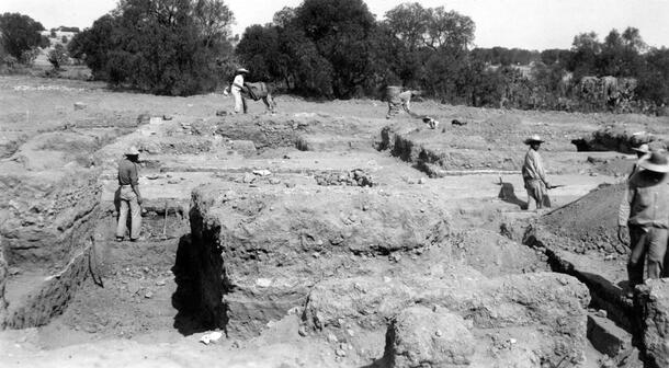 Several people wearing wide brimmed hats standing and working in various locations in a wide flat excavation site composed of partially exposed low walls. The photo caption states: "Sunken patio looking east."