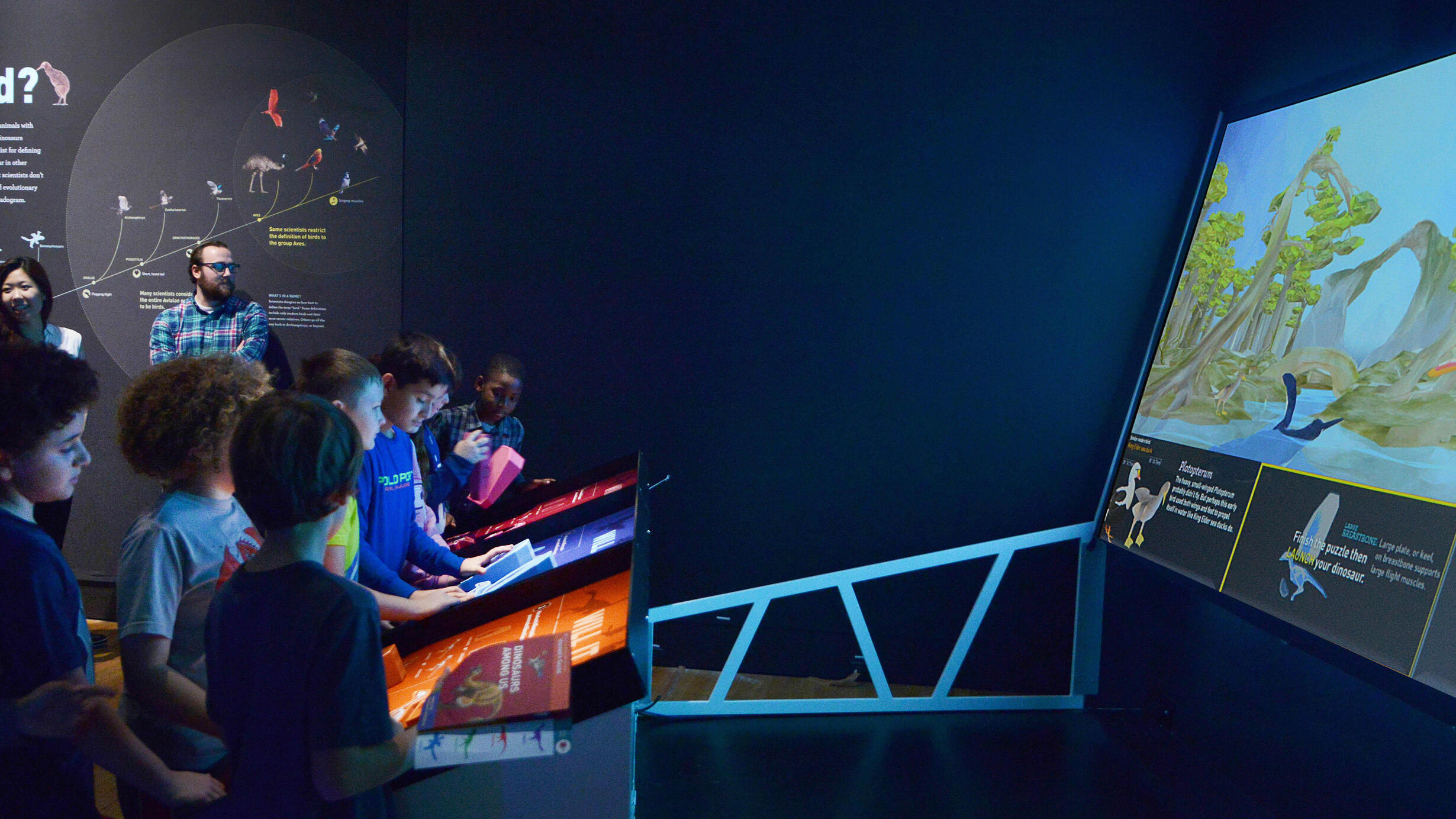 Eight children stand in front of an interactive digital display at the Dinosaurs Among Us exhibit, with two adults standing to the side.