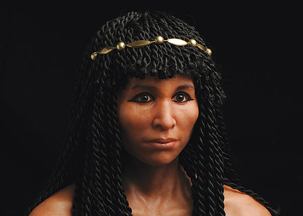 Realistic sculpture of the head and shoulders of a woman, featuring kohl-rimmed eyes, shoulder length braided hair with bangs and a gold circlet.