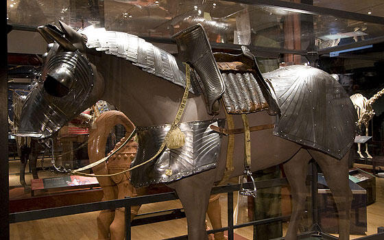 In a museum exhibition hall, a model of a horse wearing metal armor, a bridle, and a saddle with stirrups.