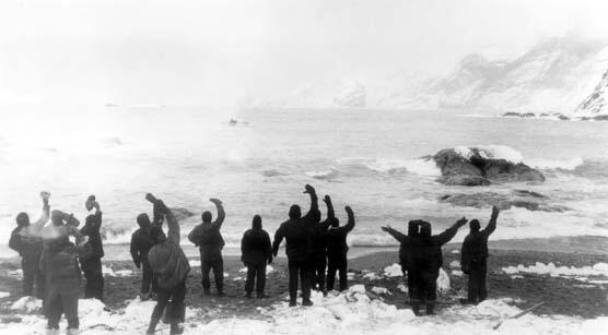 Group of 15 to 20 men on the beach of an overcast Antarctic island waving at a small ship departing in the distance.