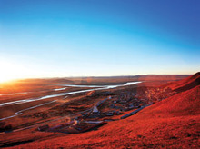 A wide shot of a blue sky over a flat landscape tinted red by the sun low in the sky, with some rolling hills, some low man-made structures, and a canal or river in the background.