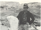 A man standing outdoors beside a massive fossil bone that is as high as the man's ribs. Dry rocky hills are in the background.