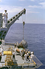 About twenty sailors stand on the deck of a ship as a space capsule is pulled from the water onto the deck via crane.