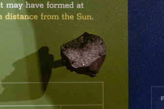 Rock specimen displayed protruding from the Museum wall, with wall text "may have formed at" and "distance from the Sun" visible above the rock.