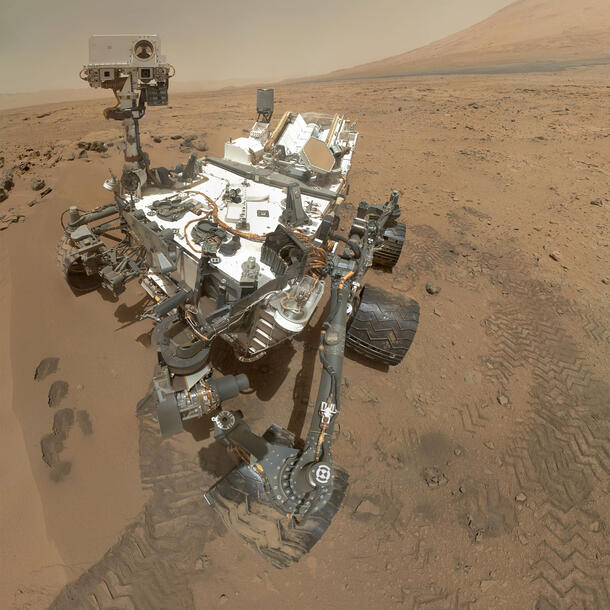 Curiosity rover, a wheeled robot on the red surface of Mars with a hill in the background. There are tracks on the ground and and a small pit
