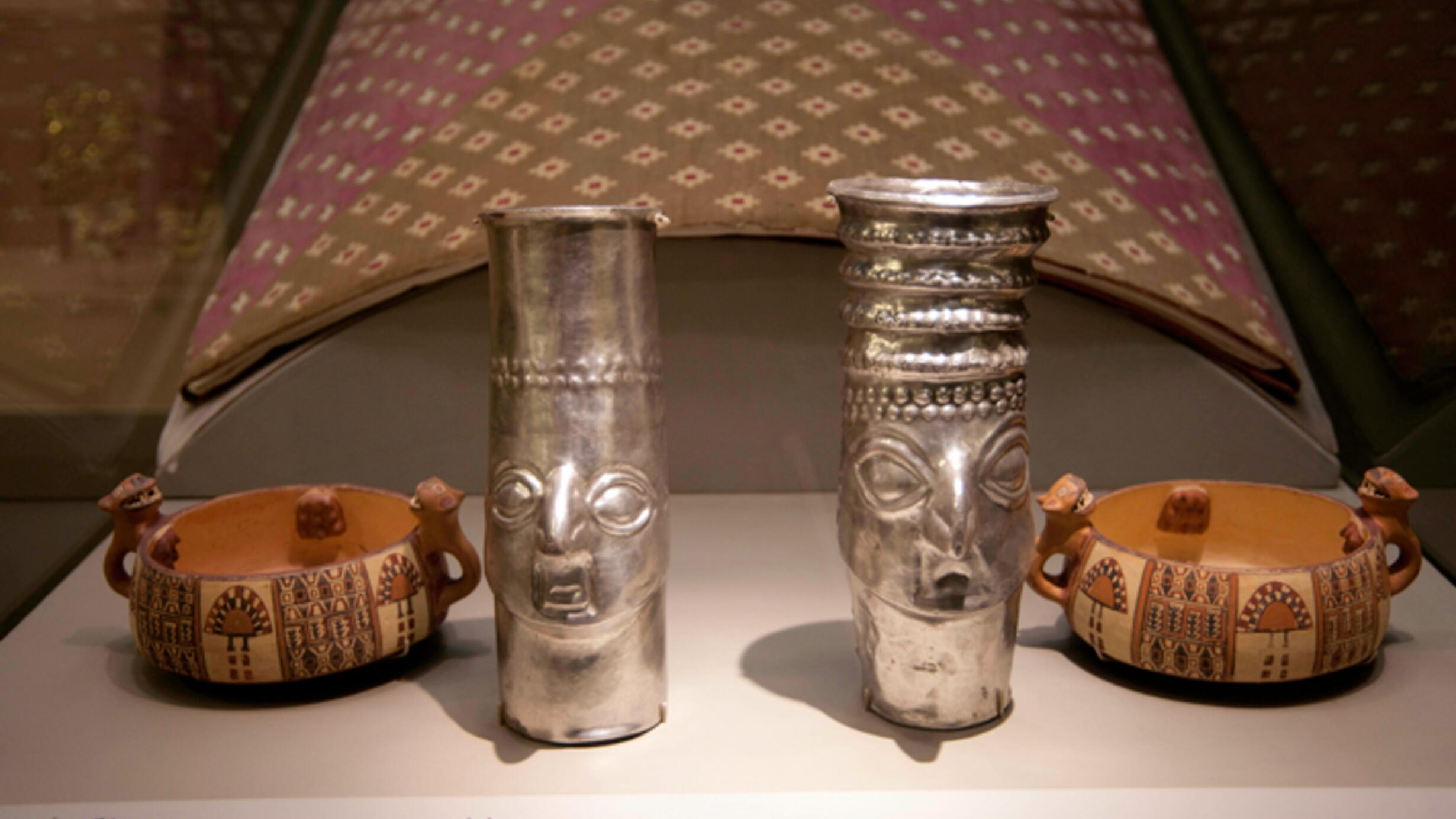 On a display table, two silver cylindric vessels decorated with face carvings and two bowls with intricately carved handles and painted designs.