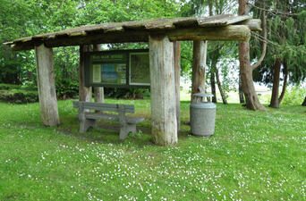 A bench and explanatory placard for Old Man House beneath a wooden T-shaped covering in a grassy field among trees.