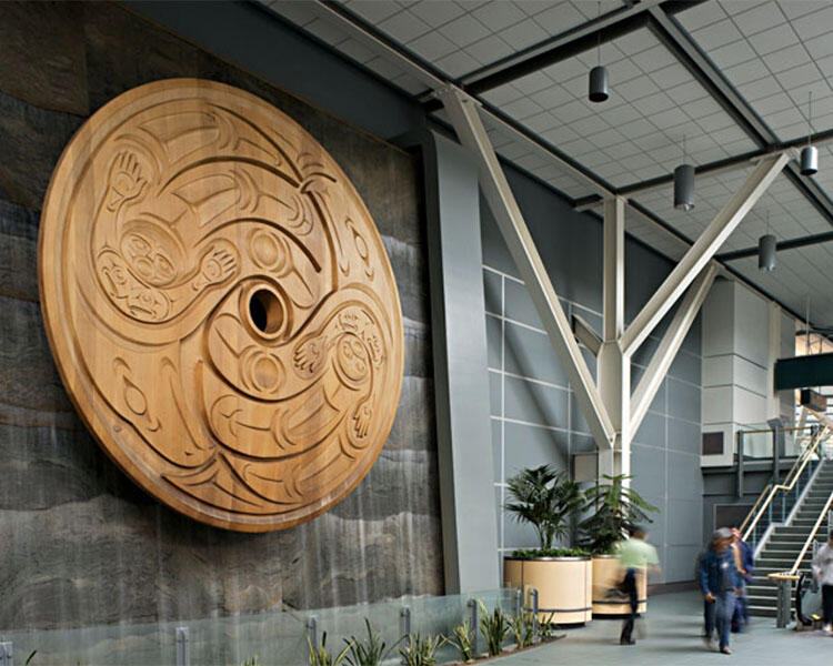 A large scale artwork—a wooden, circular form carved with fluid figures of people, a bird's face, and other abstract forms.