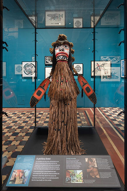 Sculpture comprised of red cedar wood and bark, synthetic hair, bear fur, seagull feather quills, acrylic paint and more, housed inside a glass display case.