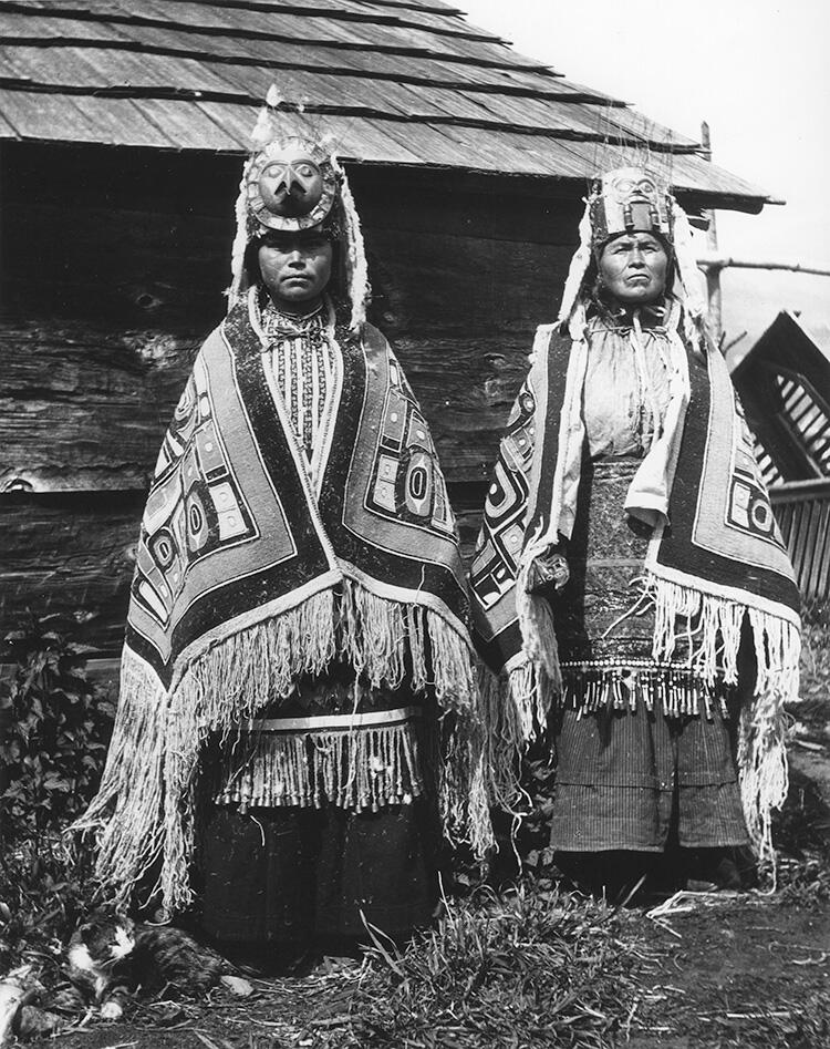 Two women standing in front of wooden building wear elaborate headdresses and thick patterned robes. 