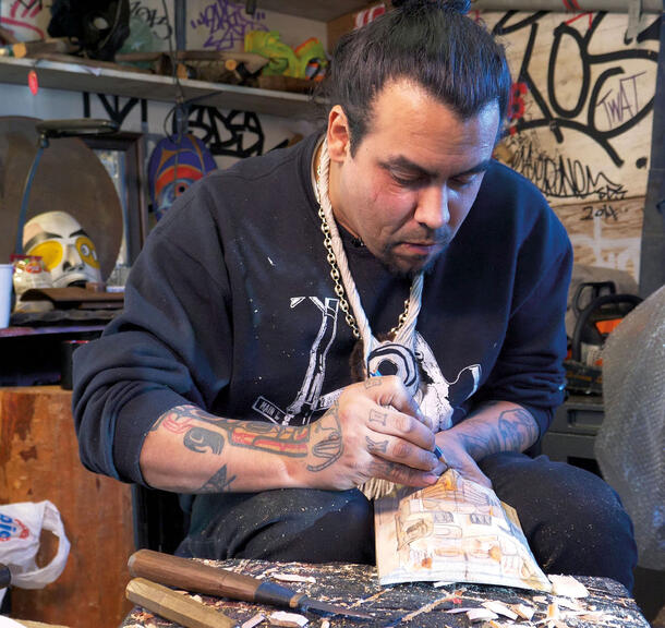 Man in a sweatshirt with tattooed arms sits in a studio making pencil markings on a carved wooden piece.