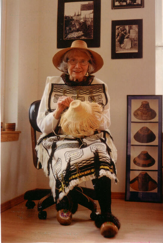 Woman in a woven hat, patterned outfit and slippers sits in a rolling chair weaving a hat. 