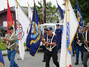 About 7 men walk down the street during parade holding various flags, one for Metlakatla and another for the U.S. Navy.