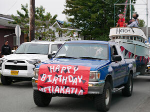 Pick-up truck with a sign reading Happy Birthday Metlakatla taped to the hood pulls a boat with 3 people on it during parade.