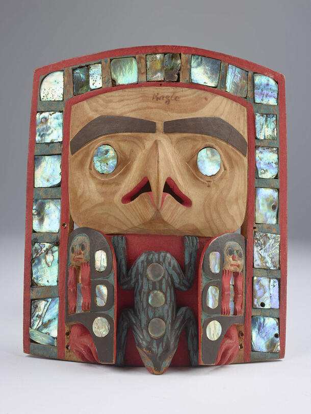 Headpiece featuring wooden carved eyes and nose, a carved frog where the mouth would be, surrounded by iridescent stone and smaller faces.