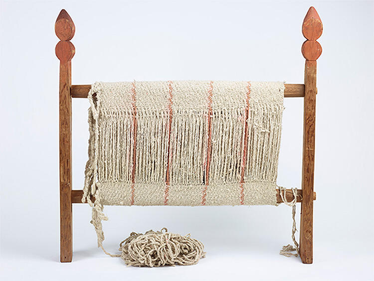 Wooden loom with two cross beams covered in partially woven piece made of white yarn with thin pink lines and ball of yarn below the loom.