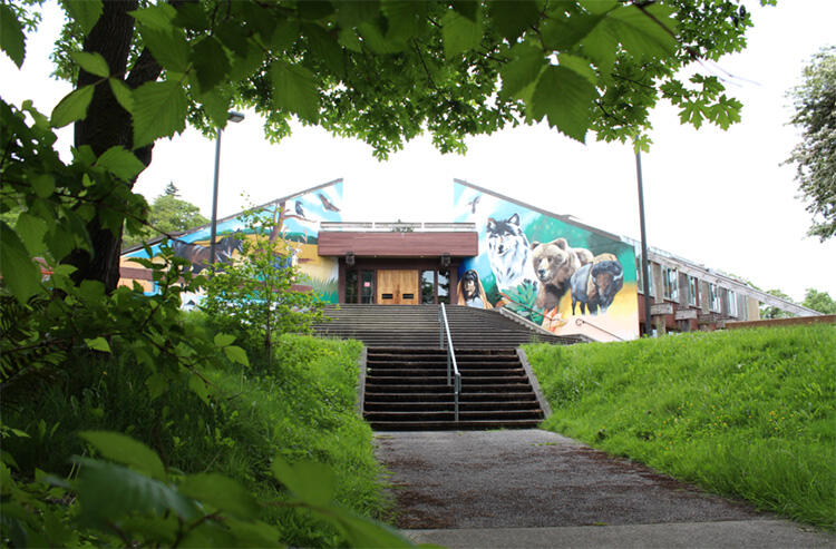 Concrete path and stairs flanked by greenery leads to front of low triangular building painted with mural of wolf, bear, buffalo, and other animals.