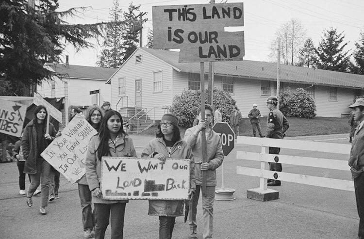 6 young people walk holding signs reading "This Land Is Our Land," "We Want Our Land Back," and cut off text "In Your Winning You Found Shame, etc." 