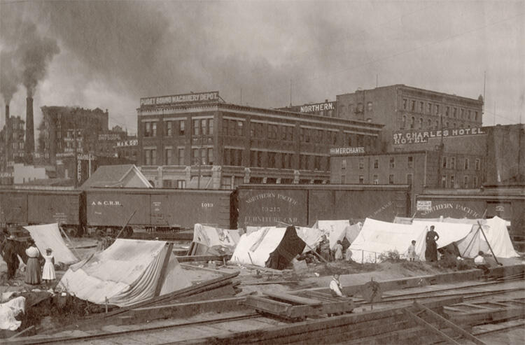 Ten white tents pitched between railroad tracks, with a train and industrial, city buildings in the background and two smoke stacks in upper left. 