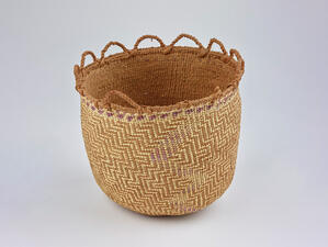 Round woven basket trimmed at the top with protruding woven half-circles in a row on one half and one small handle on the other half. 