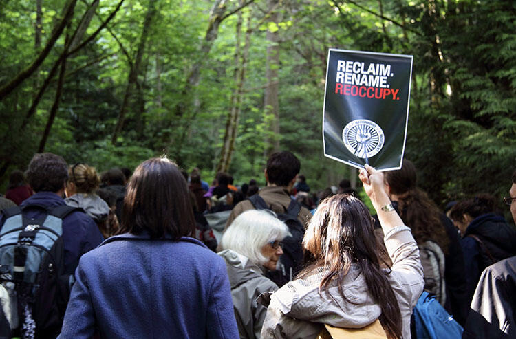 A group of people march through the woods. One holds a sign that says: Reclaim, Rename, Reoccupy.
