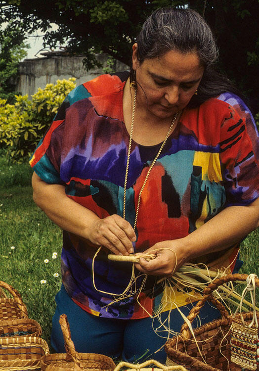  Woman in brightly patterned shirt hand-weaves part of a basket, with a number of finished woven baskets in the foreground.
