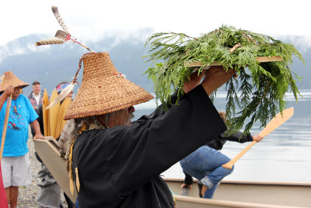 A man in a woven cedar hat lifts up a plank with cedar branches.