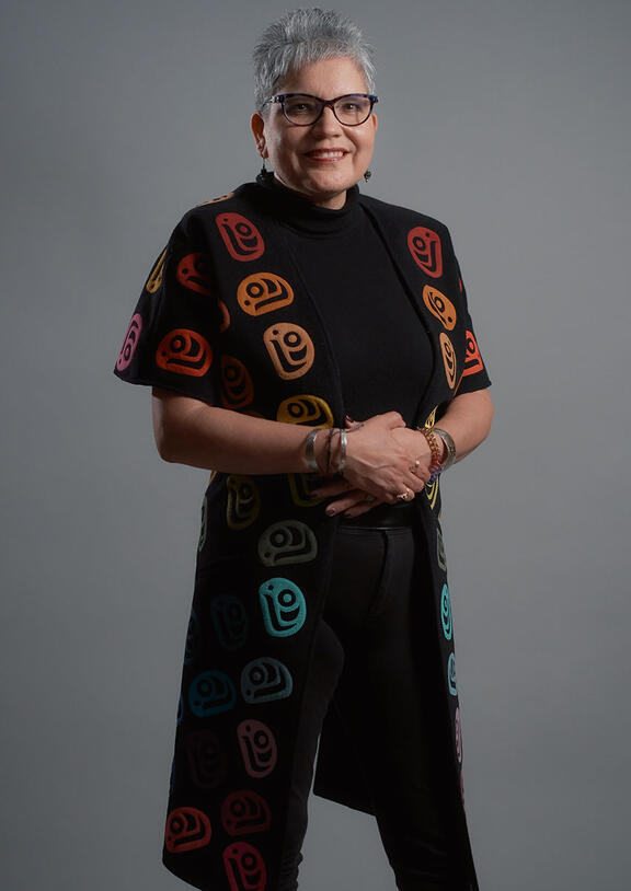 Woman with short gray hair and glasses stands with her hands held in front of her, wearing a short-sleeve jacket with multi-color patterned circles.