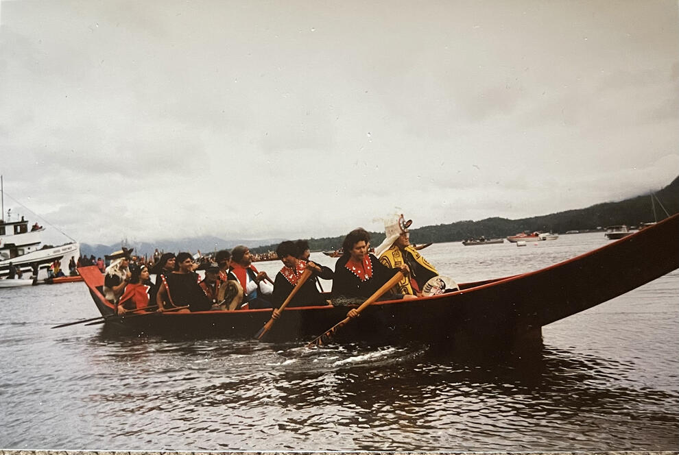 In an archival photo, a dozen people paddle a large wooden canoe.