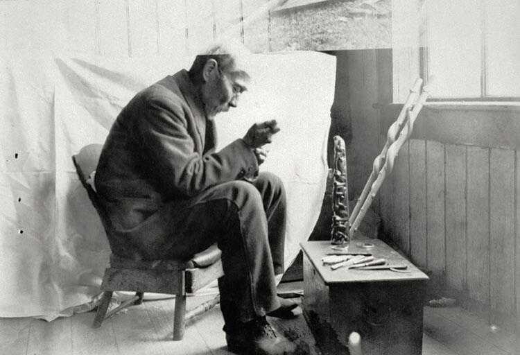 Man sitting in a small chair carving something in front of small table with a carving placed on it. 