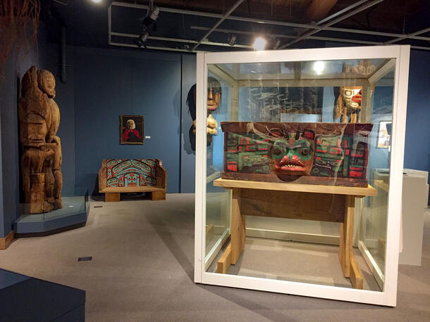 A brightly painted and decorated carved wooden chest sits in a glass cube museum display in a gallery with other wooden carvings and masks on view.