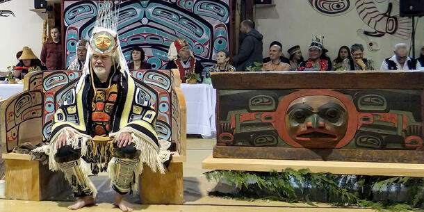 Man in headdress and patterned robe sits on painted, wooden settee with carved chest to his right and row of people seated at banquet table behind.