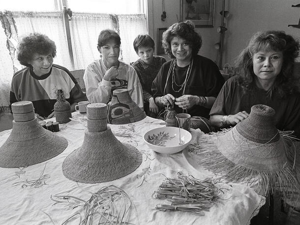 Four women and boy sit at a table indoors weaving hats.