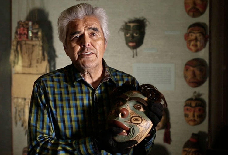 Man in patterned shirt holds a carved mask in his hands, additional masks on a gallery wall behind him.