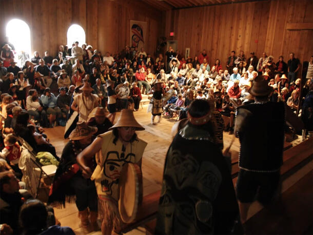 Large crowd of people in a room, including performers with drums in a circle in the center and large seated audience around three sides of performers.